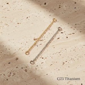 May include: Two delicate chain extenders, one gold-toned and one silver-toned, lie diagonally on a textured, beige surface. Each chain has a small loop at both ends. The text "G23 Titanium" is visible at the bottom.