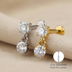May include: Two push pin labret earrings, one silver and one gold, each featuring two clear, round-cut cubic zirconia stones. The earrings are displayed on a beige fabric. The text "G23 Titanium Push Pin Labret" is visible.