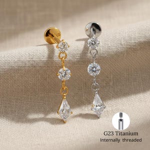 Pode incluir: Dois brincos pendurados, um dourado e outro prateado, cada um com três pedras preciosas claras e brilhantes. O brinco dourado tem uma gema em forma de gota na parte inferior. O texto "G23 Titanium Internally threaded" é visível.