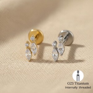 Puede incluir: Dos pares de pendientes de botón, uno dorado y otro plateado, cada uno con una pequeña gema redonda y una gema más grande con forma de marquesa. Los pendientes se muestran sobre una superficie texturizada de tono neutro. El texto dice: "G23 Titanium Internally threaded."