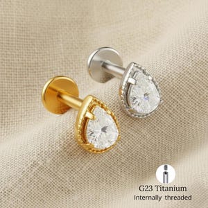 以下が含まれることがあります： G23チタン製の内部ねじ付きピアス2つ。片方は金色、もう片方は銀色です。各ピアスには、洋梨型の透明な宝石が付いています。ピアスはニュートラルカラーの生地に展示されています。