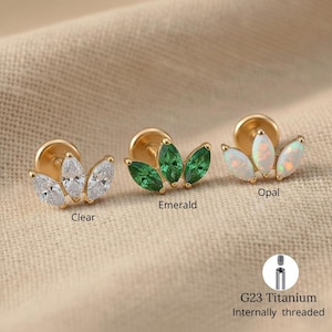 May include: Three gold-coloured, internally threaded G23 titanium earrings with a floral design. One earring features clear stones, another emerald-coloured stones, and the third opal-coloured stones. The text "Clear", "Emerald", and "Opal" are below each earring.