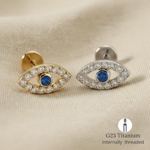 Puede incluir: Dos pendientes en forma de ojo. Uno es dorado con un centro azul y rodeado de piedras transparentes. El otro es plateado con un centro azul y rodeado de piedras transparentes. El texto "G23 Titanium Internally threaded" está debajo.