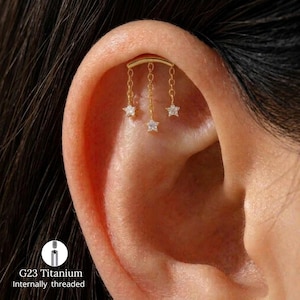 May include: A gold-toned cartilage earring featuring a curved bar with three dangling chains, each ending in a small star-shaped charm set with a clear stone. The jewellery is made of G23 titanium and features an internally threaded post design.