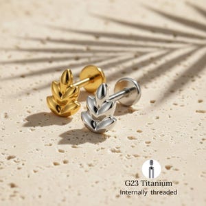 May include: Two leaf-shaped stud earrings, one gold-colored and one silver-colored, are displayed on a textured surface. The earrings have a flat back and a decorative leaf design. The text "G23 Titanium Internally threaded" is visible.