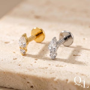 Könnte beinhalten: Zwei Ohrstecker mit birnenförmigem Design, jeweils mit einem großen, klaren, tropfenförmigen Stein über einem kleineren, runden Stein. Ein Ohrring ist goldfarben, der andere silberfarben, beide mit flacher Rückseite und Stecker.