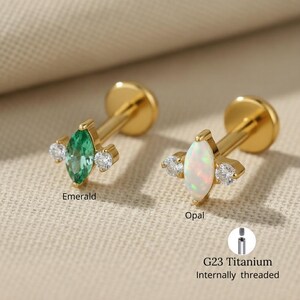Può includere: Due orecchini in titanio G23 color oro, filettati internamente. Uno presenta una pietra centrale di smeraldo taglio marquise, affiancata da due pietre chiare più piccole. L'altro ha una pietra centrale di opale, anch'esso con due pietre chiare. Il testo "Emerald" e "Opal" è visibile.