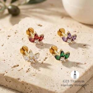 Puede incluir: Cuatro pendientes de titanio G23 dorados con rosca interna. Cada pendiente presenta un diseño floral con piedras preciosas de colores: rojo, morado, blanco y verde. Una pequeña gema transparente está en el centro de cada diseño floral.