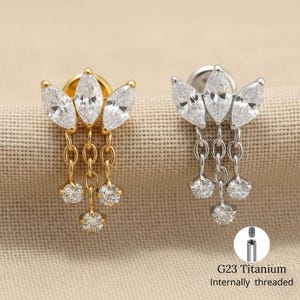 May include: Two stud earrings, one gold-toned and one silver-toned, each with a floral design of three marquise-cut clear stones. Each earring has three chains with round, clear stones. The text "G23 Titanium Internally threaded" is also visible.