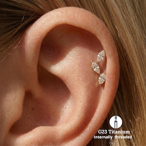 Peut inclure: Un piercing de cartilage d'oreille doré composé de trois pierres précieuses transparentes taille marquise disposées en grappe verticale. Le bijou est en titane G23 et est fileté intérieurement, comme indiqué par le texte et le schéma.