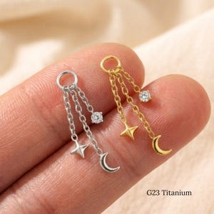 May include: Two G23 titanium jewelry charms, one silver-toned and one gold-toned. Each charm features a circular ring attached to three delicate chains of varying lengths, ending in a small clear gemstone, a four-pointed star, and a crescent moon.