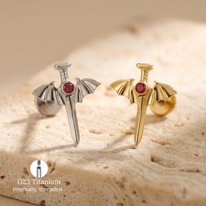 May include: Two stud earrings, one silver and one gold, each featuring a sword design with bat-wing accents and a red gemstone. The earrings are made of G23 titanium and are internally threaded.