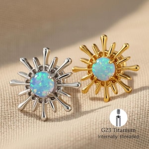 Puede incluir: Dos pendientes en forma de sol con una ópalo azul claro en el centro. Un pendiente es plateado, el otro dorado. Los pendientes tienen un diseño puntiagudo alrededor del ópalo. También es visible el texto "G23 Titanium Internally threaded".