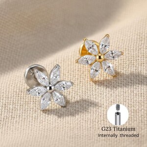 May include: Two flower-shaped earrings. One is silver, the other gold. Each earring has six clear, marquise-cut stones around a central bead. The image includes a close-up of the earring post and the text "G23 Titanium Internally threaded."