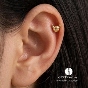 May include: A gold-colored ear stud shaped like a small devil face with two horns. The jewelry is positioned in the upper ear cartilage. A small circular inset shows the G23 titanium internally threaded post used for this piece of body jewelry.