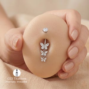 Può includere: Un piercing all'ombelico argentato con un design a farfalla pendente. La gemma superiore è una pietra trasparente e rotonda. Il testo "G23 Titanium Internally threaded" è visibile.