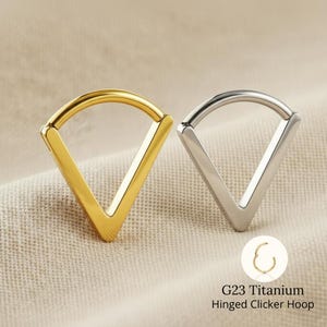 Può includere: Due orecchini a cerchio incernierati, uno dorato e uno argentato, a forma di V. Gli orecchini sono in titanio G23. Il testo "G23 Titanium Hinged Clicker Hoop" è in basso a destra.