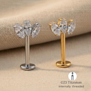 May include: Two labret studs with a cluster of three clear marquise-cut cubic zirconia stones. One stud is silver-colored, the other is gold-colored. The base of each stud is round. The text "G23 Titanium Internally threaded" is visible.