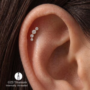 Peut inclure: Un bijou de cartilage d'oreille doré composé d'une rangée incurvée de quatre pierres rondes transparentes de tailles graduées. Le bijou est en titane G23 à filetage interne. Le design suit la courbe naturelle du cartilage de l'oreille.