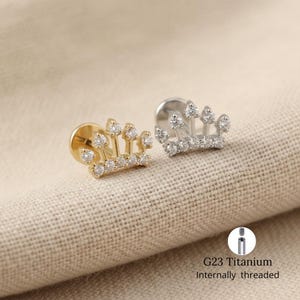 Puede incluir: Dos pendientes en forma de corona, uno dorado y otro plateado, cada uno adornado con pequeñas piedras preciosas transparentes. Los pendientes se muestran sobre una superficie texturizada de color neutro. El texto "G23 Titanium Internally threaded" también es visible.