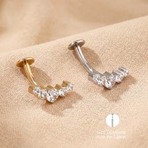 Könnte beinhalten: Zwei gebogene Labret-Piercings, eines goldfarben und eines silberfarben, jeweils mit einer Reihe klarer, funkelnder Edelsteine. Die Piercings haben eine flache, kreisförmige Rückseite und einen gebogenen Stab. Der Text "G23 Titanium Push Pin Labret" ist ebenfalls enthalten.