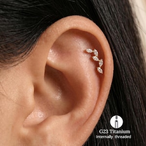 Peut inclure: Un piercing d'oreille doré avec six pierres précieuses claires de taille marquise disposées en ligne incurvée. Le piercing est situé sur le cartilage supérieur de l'oreille. Le texte "G23 Titanium Internally threaded" est visible.