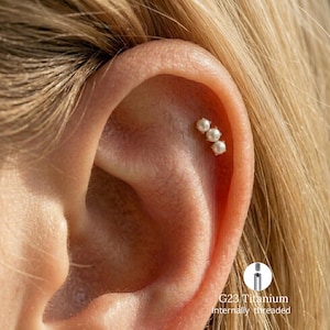 May include: A cartilage earring featuring three small, round white pearls arranged in a vertical row. The jewelry is internally threaded and crafted from G23 titanium. The earring is positioned in the upper ear cartilage, showcasing a metallic finish.