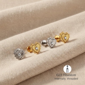 May include: Four heart-shaped stud earrings in gold and silver tones, each with a central clear gem. The earrings are arranged on a beige fabric. The text "G23 Titanium Internally threaded" is visible.