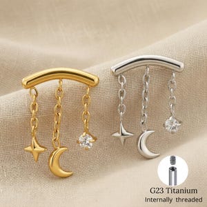 Può includere: Due orecchini a tema celeste. Uno è dorato, l'altro argentato. Ognuno presenta una barra curva con catene pendenti che tengono una stella, una luna crescente e una gemma trasparente. Il testo "G23 Titanium Internally threaded" è visibile.