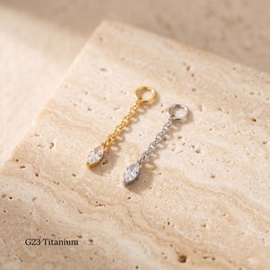 May include: Two dangling earrings, one gold-toned and one silver-toned, each featuring a chain and a teardrop-shaped, clear gemstone. The earrings are displayed on a textured, light-colored surface. The text "G23 Titanium" is visible.