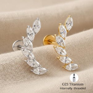 Puede incluir: Dos pendientes de perforación, uno plateado y otro dorado, cada uno con una fila curva de piedras preciosas transparentes con corte marquesa. Los pendientes se muestran sobre un fondo de tela neutro. El texto "G23 Titanium Internally threaded" es visible.