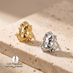 May include: Two frog-shaped body jewellery pieces. One is gold, the other silver. Both have a textured surface with small, round accents. The text "G23 Titanium Internally threaded" is visible.