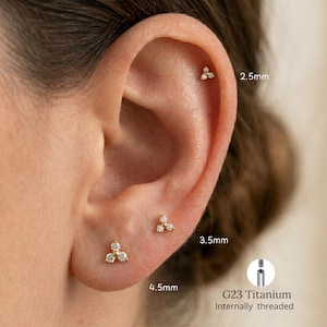 Peut inclure: Gros plan sur une oreille ornée de trois petites boucles d'oreilles dorées, chacune avec un groupe de trois pierres précieuses transparentes. Les boucles d'oreilles sont étiquetées avec des mesures : 2,5 mm, 3,5 mm et 4,5 mm. L'image comprend également un schéma d'une tige de boucle d'oreille filetée interne en titane G23.