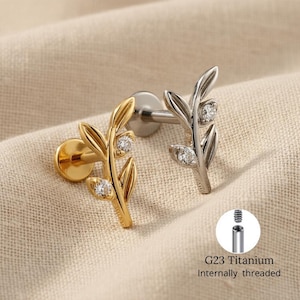 Può includere: Due labret a forma di foglia. Uno è dorato, l'altro argentato. Ognuno presenta tre gemme rotonde e trasparenti. I labret sono su uno sfondo di tessuto beige. Il testo "G23 Titanium Internally threaded" è in un cerchio.