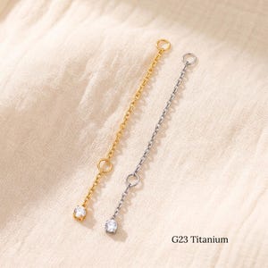 May include: Two G23 titanium jewelry chain attachments, one gold-toned and one silver-toned. Each features a delicate metal link chain connected to a small circular jump ring at the top and a single clear, round-cut gemstone charm at the bottom.