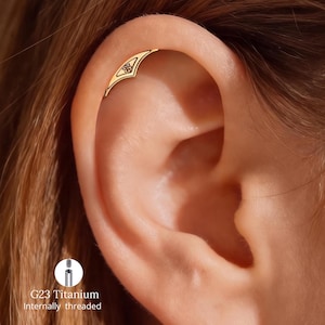 May include: A gold-colored ear cartilage piercing featuring a curved, pointed shield design with an engraved floral motif. The jewelry is G23 titanium and internally threaded, as indicated by the text and diagram in the lower left corner.