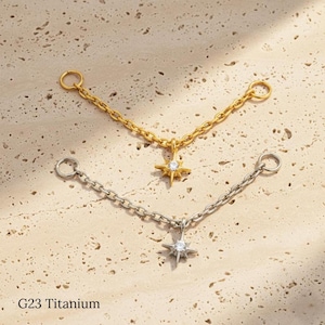 May include: Two star-shaped body jewelry pieces. One is gold-colored, the other silver-colored. Each has a chain with a ring on each end and a star pendant with a central gem. The text "G23 Titanium" is at the bottom.