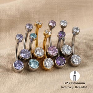 Può includere: Una collezione di piercing per ombelico curvi con pietre preziose di vari colori. Gli anelli hanno montanti in metallo argentato e dorato. Le pietre preziose sono nelle tonalità di blu, viola, rosa e trasparente. Il testo "G23 Titanium Internally threaded" è visibile.