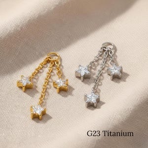 May include: Two pairs of star-shaped earrings. One pair is gold-colored, and the other is silver-colored. Each earring has three dangling stars with clear, sparkling accents. The text "G23 Titanium" is visible.