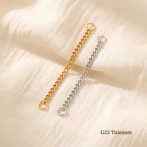 Könnte beinhalten: Zwei kurze Panzerketten, eine goldfarbene und eine silberfarbene, jeweils mit einem kleinen runden Biegering an beiden Enden. Der Text G23 Titanium ist in der unteren rechten Ecke des Bildes aufgedruckt und gibt das Material an.