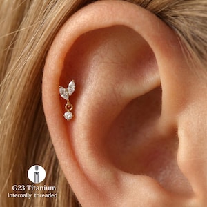 May include: A gold-colored ear piercing with a dangling design. The piercing features two marquise-cut clear gemstones and a round clear gemstone. The text "G23 Titanium Internally threaded" is also visible.