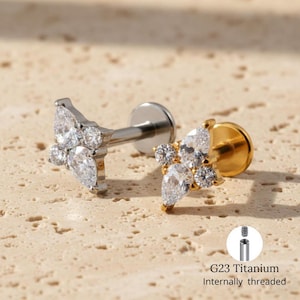 May include: Two pieces of jewellery, one silver and one gold, each featuring a cluster of clear, teardrop-shaped and round gemstones. The jewellery is designed for body piercing. The text "G23 Titanium Internally threaded" is visible.