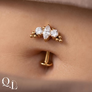 May include: A gold belly button ring with a cluster of clear, round gemstones and a central marquise-cut stone. The ring has a curved design with small gold beads on each side of the gemstones.
