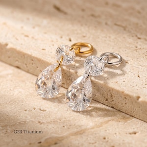May include: Two clear, teardrop-shaped earrings with a sparkling, faceted design. One earring has a gold-colored hoop and the other has a silver-colored hoop. The earrings are suspended from a small, round, clear stone. The text "G23 Titanium" is at the bottom.