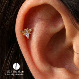 Peut inclure: Une boucle d'oreille de cartilage en forme d'abeille dorée avec une pierre de zircone cubique transparente en forme de poire pour le corps et de petites ailes. Le bijou est marqué G23 Titanium et fileté intérieurement, porté au cartilage.
