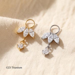 May include: Two G23 titanium body jewellery charms, one gold-toned and one silver-toned. Each charm features a circular ring at the top, three marquise-cut clear crystals arranged in a fan shape, and a single round-cut clear crystal dangling below.