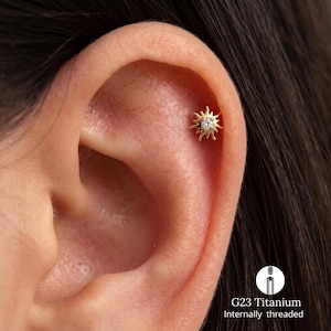 May include: A gold-colored sun-shaped ear stud featuring a central clear gemstone. The jewelry piece has radiating rays and is shown inserted into the upper ear cartilage. Text on the image indicates G23 Titanium and internally threaded.
