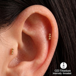 Könnte beinhalten: Zwei goldfarbene Ohrpiercings, die jeweils aus einer vertikalen Reihe von drei kleinen Kugeln bestehen. Der Schmuck ist aus G23-Titan gefertigt und verfügt über ein Innengewinde. Ein Piercing sitzt am Tragus, das andere am oberen Knorpel.