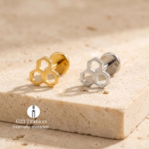 Puede incluir: Dos pendientes de botón con diseño de panal. Un pendiente es de color dorado y el otro plateado. Los pendientes son de titanio G23 y tienen rosca interna. Los pendientes se muestran sobre una superficie clara y texturizada.