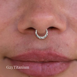 Peut inclure: Gros plan sur un anneau de septum argenté orné d'un motif complexe. L'anneau, en titane G23, est porté sur le nez d'une personne. Il a une forme incurvée en croissant avec un design détaillé.